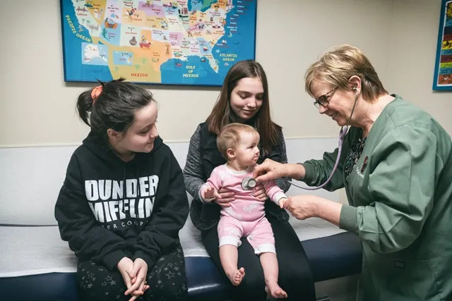 a toddler sitting on her sisters lap while a pediatirican listens to her heart